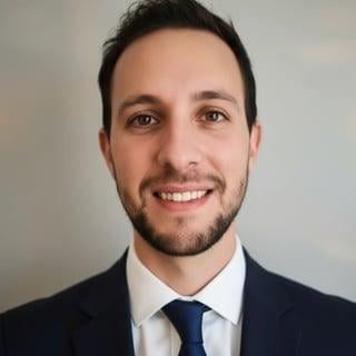 Professional headshot of a smiling man with dark hair wearing a dark suit, white dress shirt, and navy tie