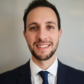 Professional headshot of a smiling man with dark hair and beard wearing a navy suit and blue tie